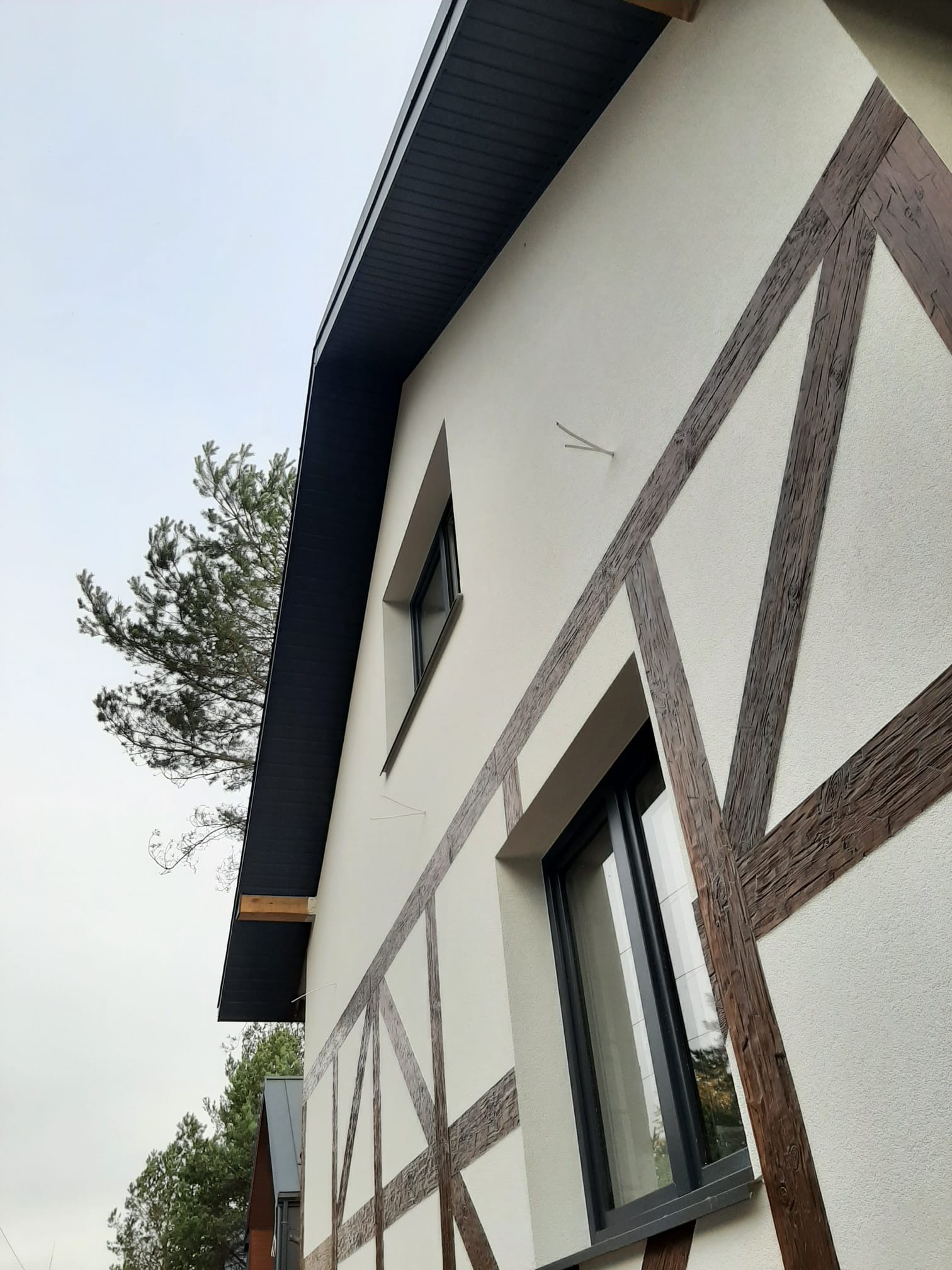 Nowoczesny dom z elewacją w stylu muru pruskiego, widok z dołu na fragment ściany z oknami i dekoracyjnymi, drewnopodobnymi belkami na jasnym tynku.