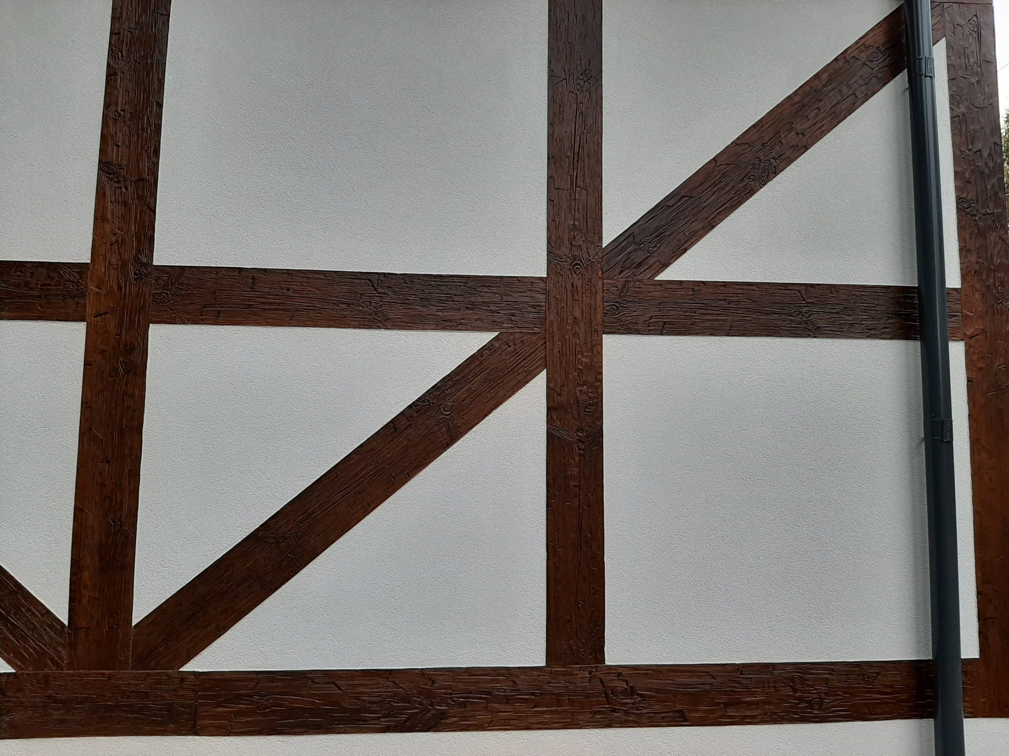 Fragment elewacji budynku z białą ścianą i dekoracyjnymi, ciemnymi belkami imitującymi konstrukcję szkieletową, z widoczną rurą spustową po prawej stronie.