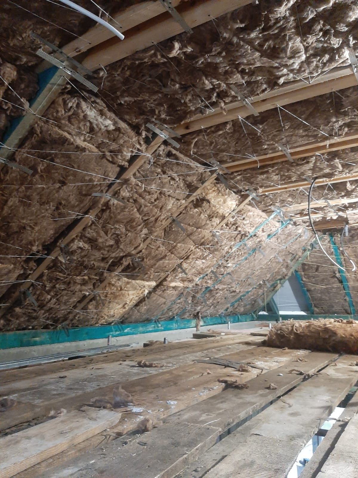 Poddasze w trakcie ocieplania wełną mineralną, widoczne drewniane krokwie i stalowe elementy mocujące. Nieukończona podłoga z desek.