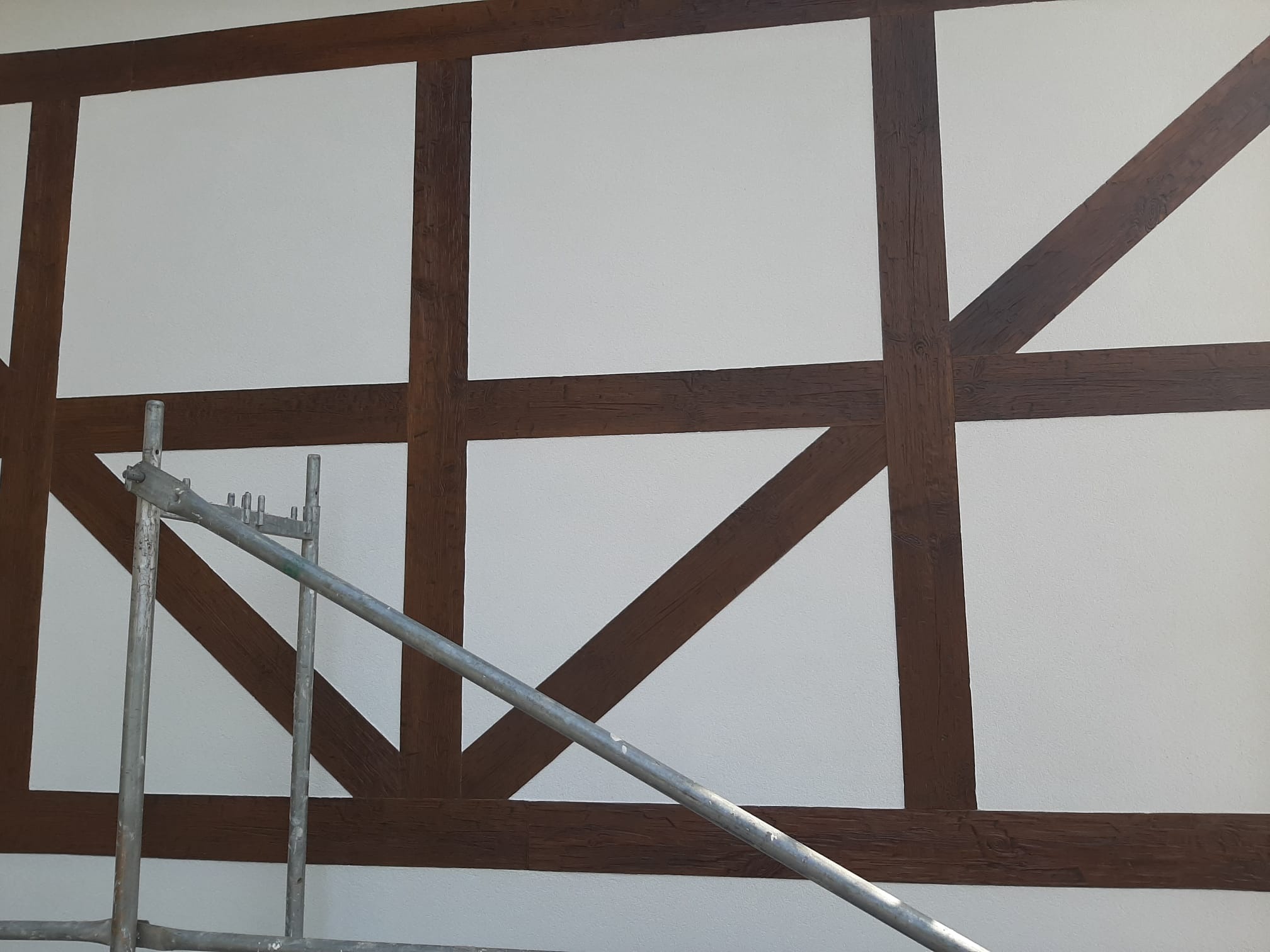 Biała ściana z dekoracyjną konstrukcją z ciemnego drewna przypominającą mur pruski, częściowo zasłonięta przez metalowe rusztowanie.