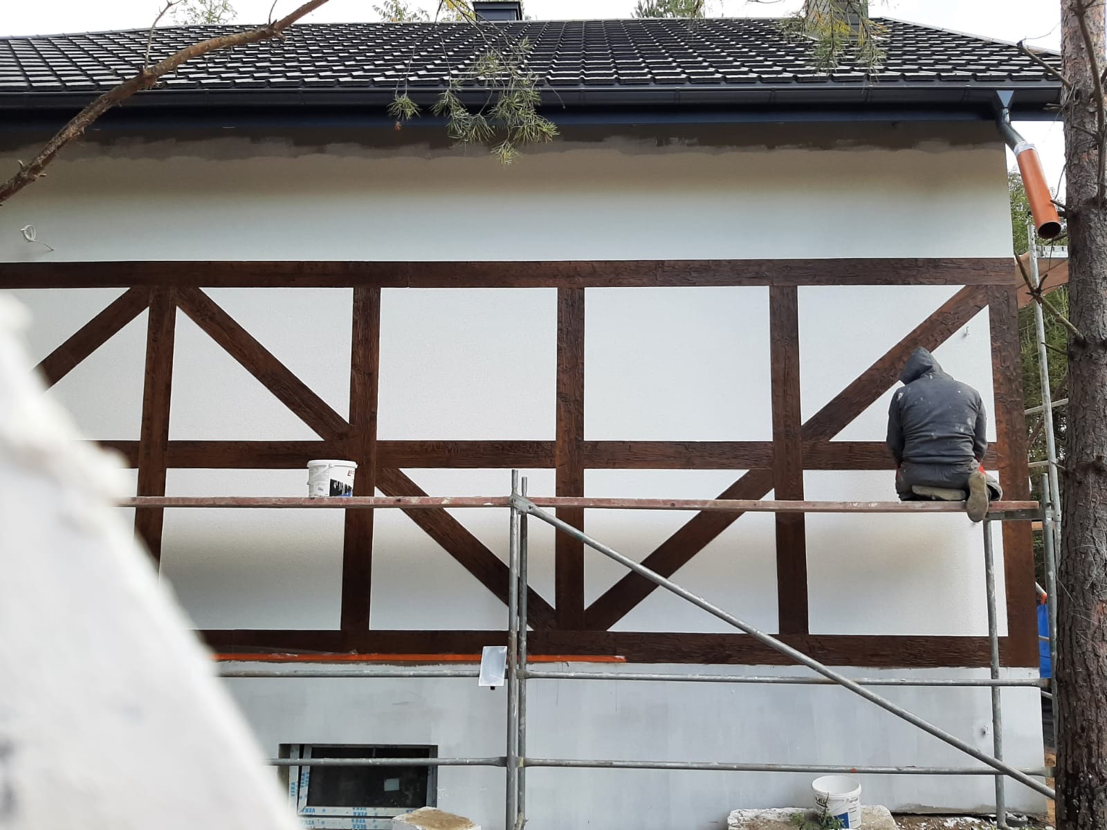 Pracownik siedzi na rusztowaniu podczas malowania elewacji budynku z elementami drewnianej konstrukcji szkieletowej, widoczne puszki z farbą.