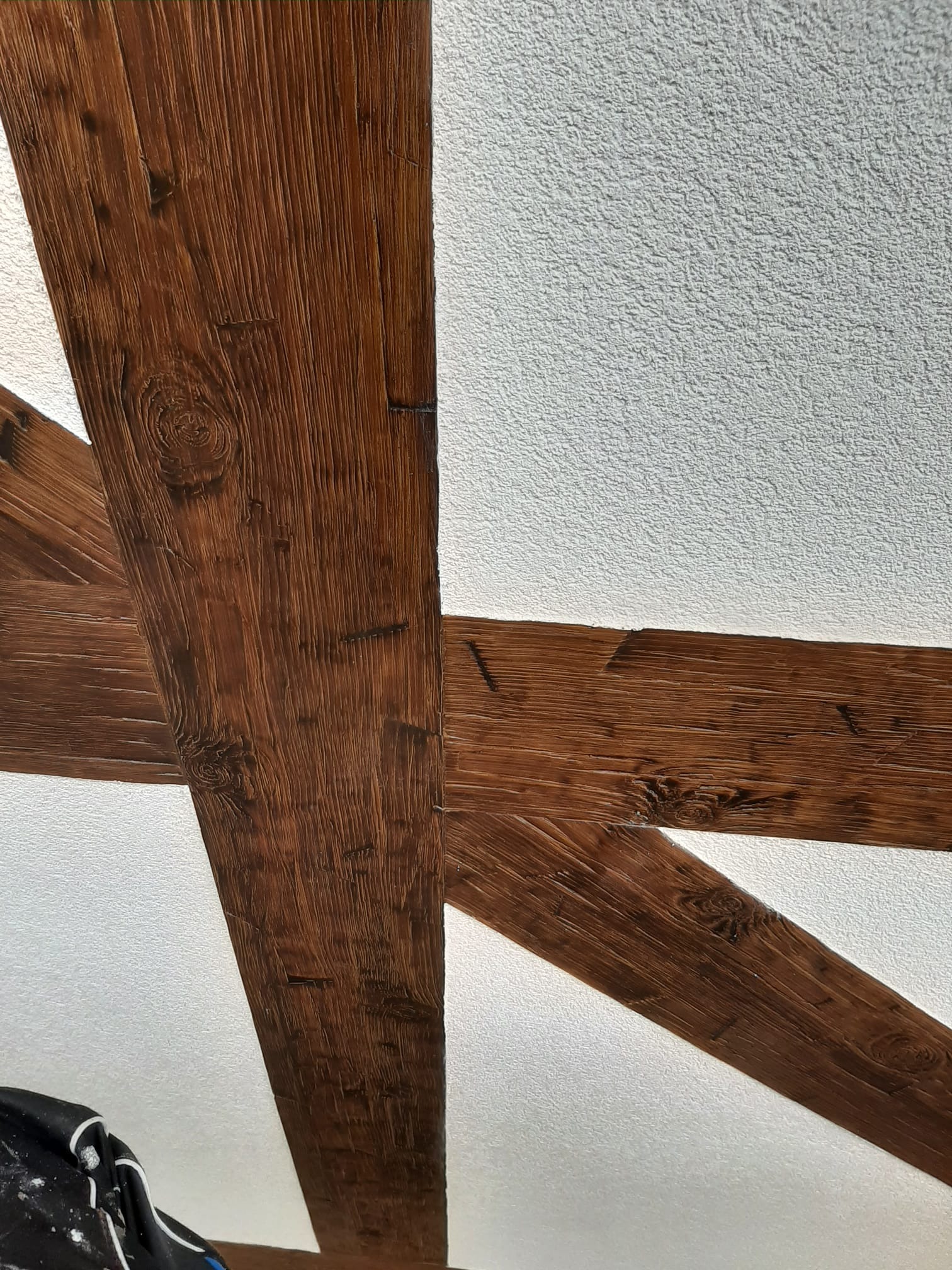 Drewniane belki stropowe w rustykalnym stylu na tle białego, teksturowanego sufitu. Widoczny fragment ubrania roboczego w rogu kadru.