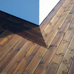 Fragment tarasu z desek kompozytowych w dwóch odcieniach brązu, przylegającego do białego narożnika budynku, widoczne wyraźne usłojenie drewna.