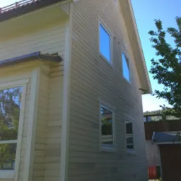 Świeżo pomalowany, dwukondygnacyjny dom z jasną elewacją drewnianą, widoczne okna z białymi ramami i fragment dachu z metalową barierką, całość na tle błękitnego nieba.