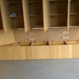 Jasne drewniane szafki kuchenne z otwieranymi klapami i g&oacute;rnymi szafkami, widok na okno z widokiem na zewnątrz, minimalistyczny design.