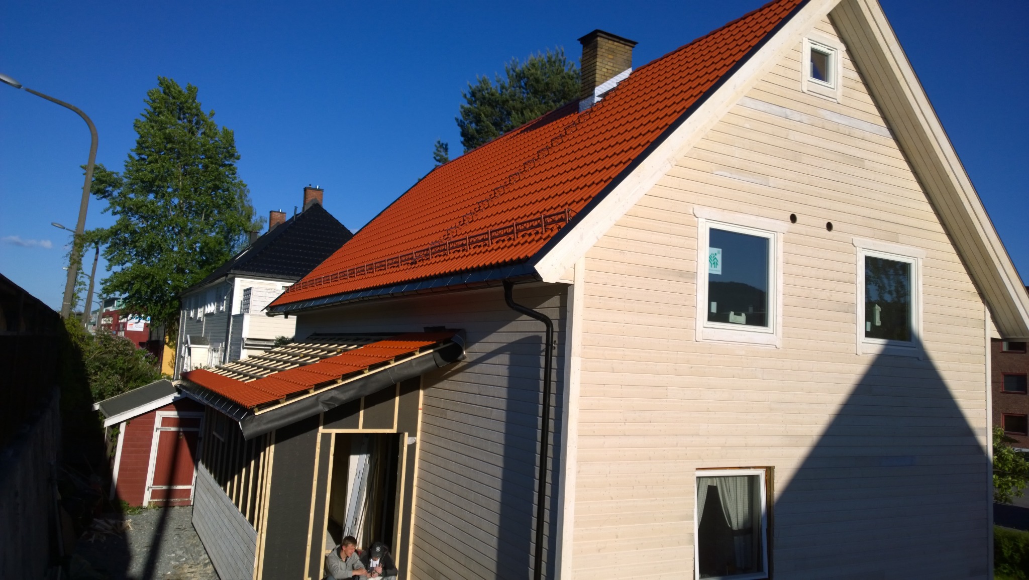 Dom w trakcie remontu elewacji i dachu: częściowo pomalowana elewacja na jasno, nowy dach z czerwonej dachówki, widoczni dwaj pracownicy w otworze okiennym dobudówki.