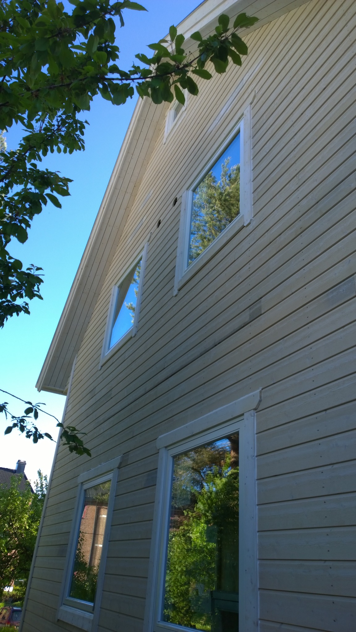 Elewacja domu z drewnianą boazerią pomalowaną na jasny kolor, widok z dołu, w oknach odbija się zieleń drzew, Wlewsk.