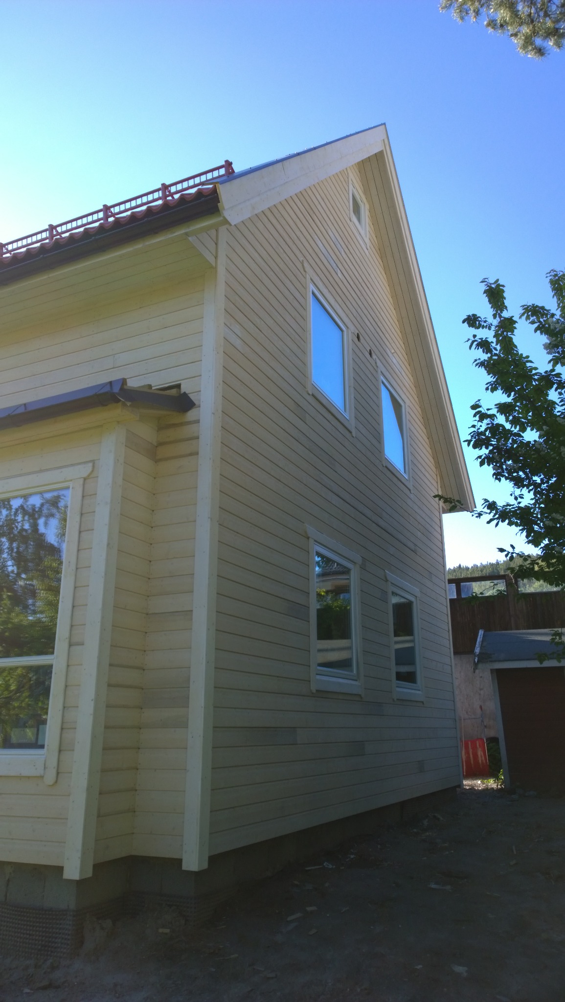 Świeżo pomalowany, dwukondygnacyjny dom z jasną elewacją drewnianą, widoczne okna z białymi ramami i fragment dachu z metalową barierką, całość na tle błękitnego nieba.