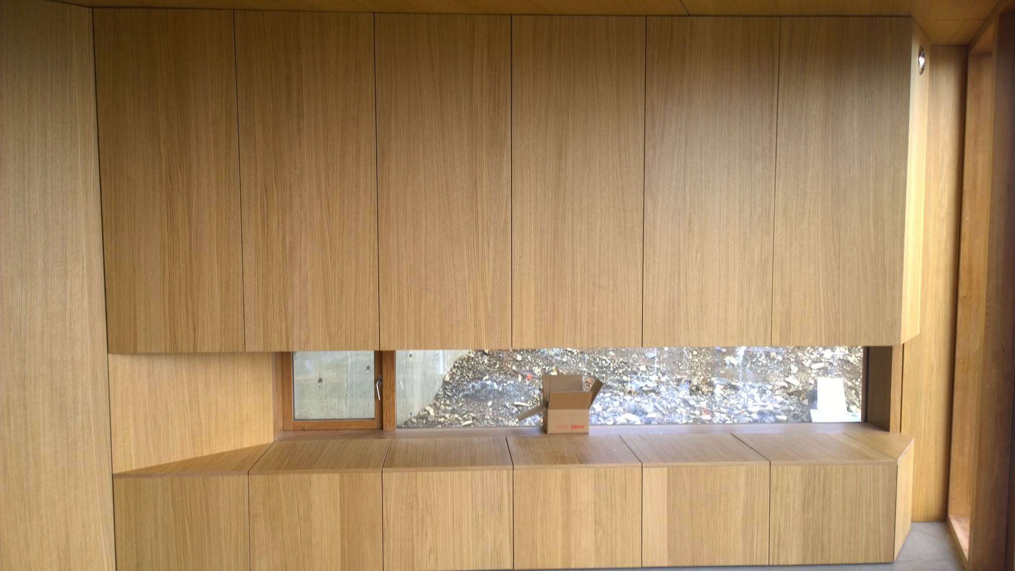 Drewniana zabudowa meblowa z szafkami wiszącymi i dolnymi, tworząca jednolitą powierzchnię z oknem i kartonowym pudełkiem na blacie.