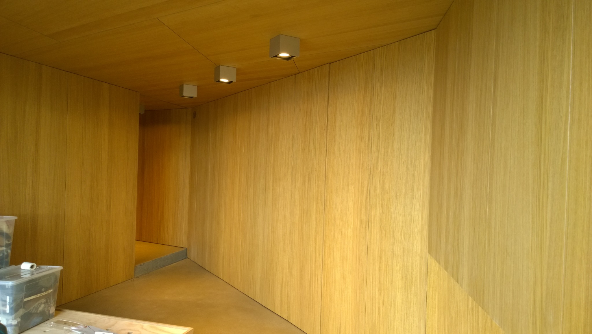 Wnętrze wykończone panelami drewnianymi w odcieniach beżu i brązu, z trzema kwadratowymi lampami sufitowymi i widocznym przejściem do innego pomieszczenia, z plastikowymi pojemnikami w lewym dolnym...