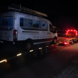 Biały bus firmy Solar Plus z drabiną na dachu, przewożony na ciemnej lawecie oświetlonej czerwonymi światłami w nocy. Na busie reklama usług czyszczenia paneli fotowoltaicznych.