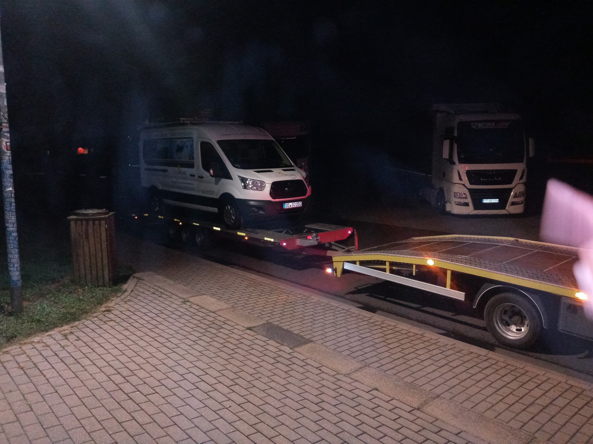 Nocne ujęcie lawety z białym busem na platformie, obok stoi biała ciężarówka z napisem 'SZWEDEK' nad przednią szybą.