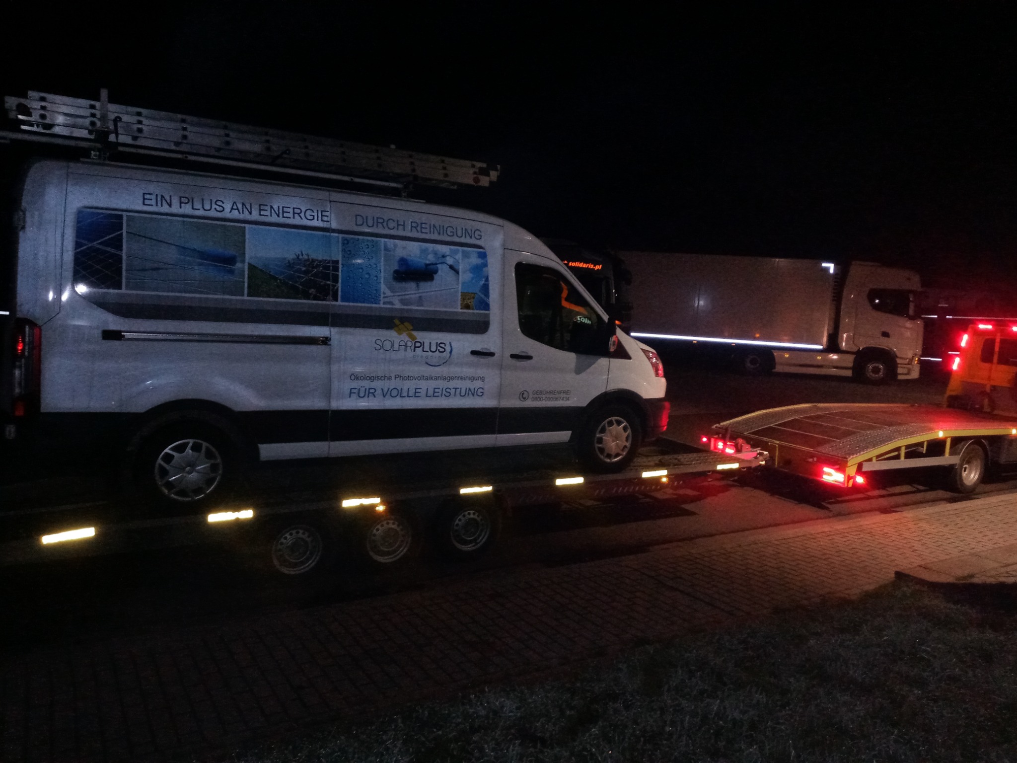 Laweta przewozi busa firmy Solarplus z drabiną na dachu; w tle zaparkowana ciężarówka; nocne oświetlenie