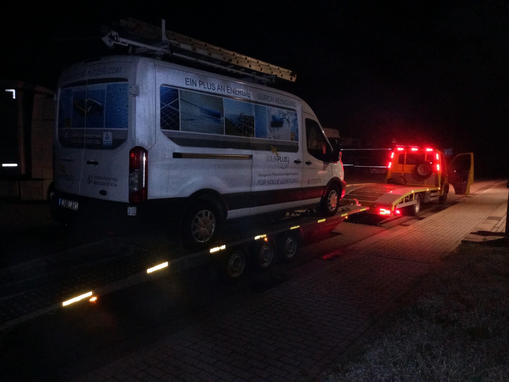 Biały bus firmy Solar Plus z drabiną na dachu, przewożony na ciemnej lawecie oświetlonej czerwonymi światłami w nocy. Na busie reklama usług czyszczenia paneli fotowoltaicznych.