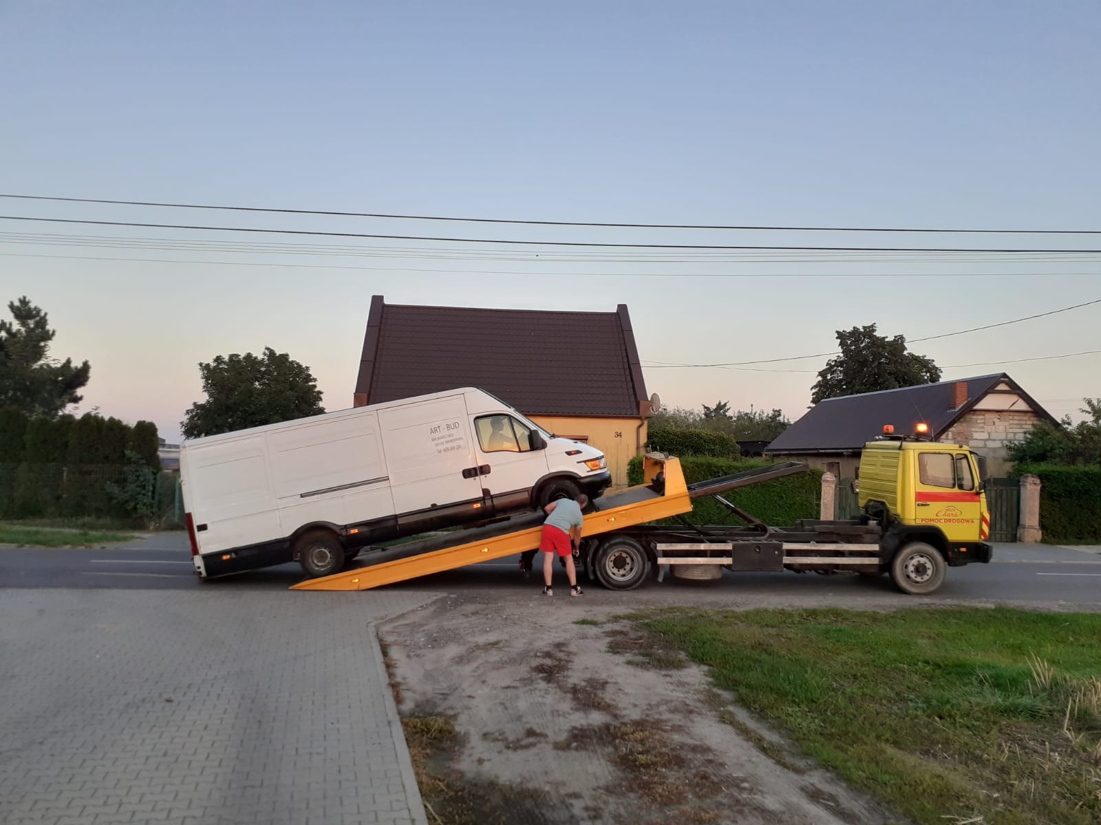 Żółta laweta z białym busem na platformie, mężczyzna w czerwonych spodenkach przy kole pojazdu, w tle domy i drzewa, zdjęcie zrobione o zmierzchu.