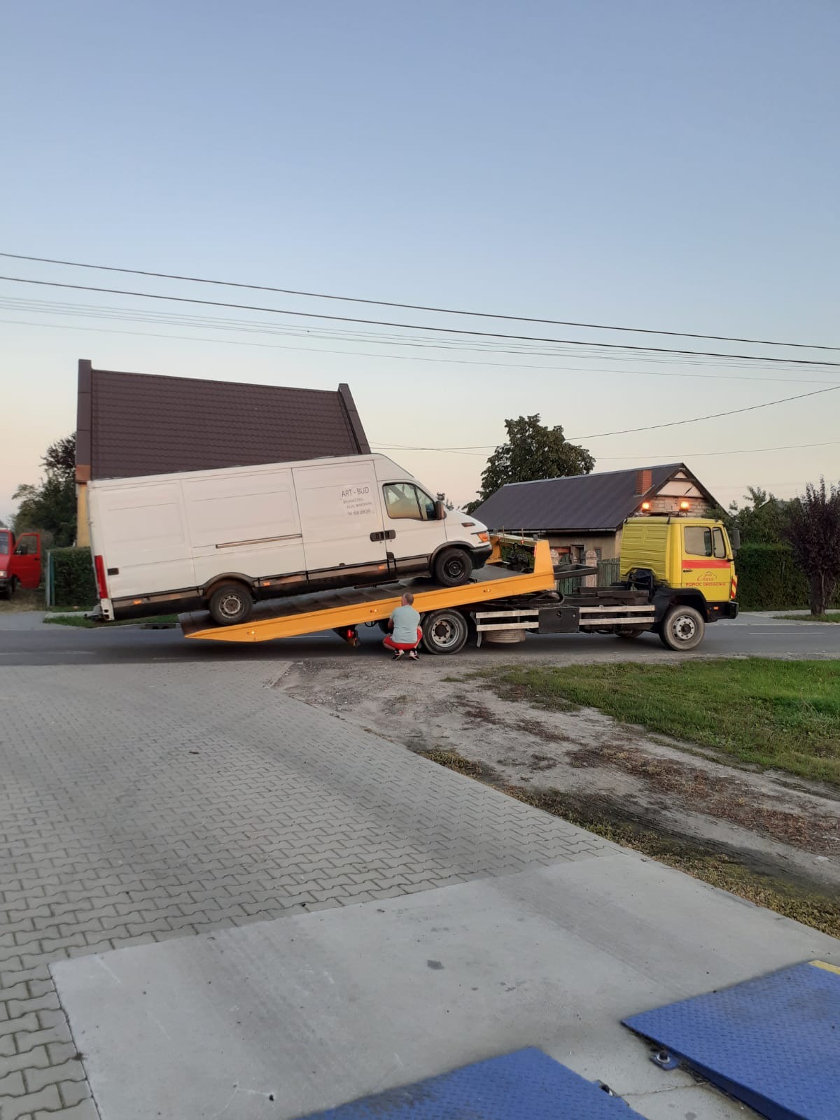 Żółta ciężarówka pomocy drogowej z białym busem na platformie; mężczyzna w czerwonej koszulce kuca przy tylnym kole. Dom z brązowym dachem w tle.