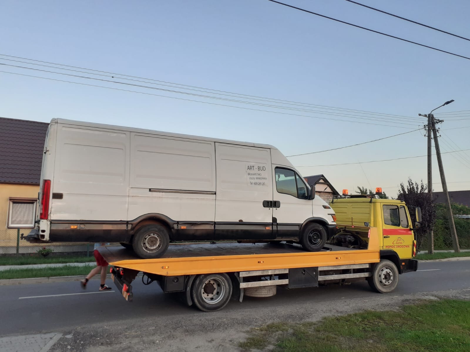 Żółta laweta z białym busem firmy ART-BUD na platformie, widziana z boku na tle zabudowań i słupa energetycznego, w tle błękitne niebo.