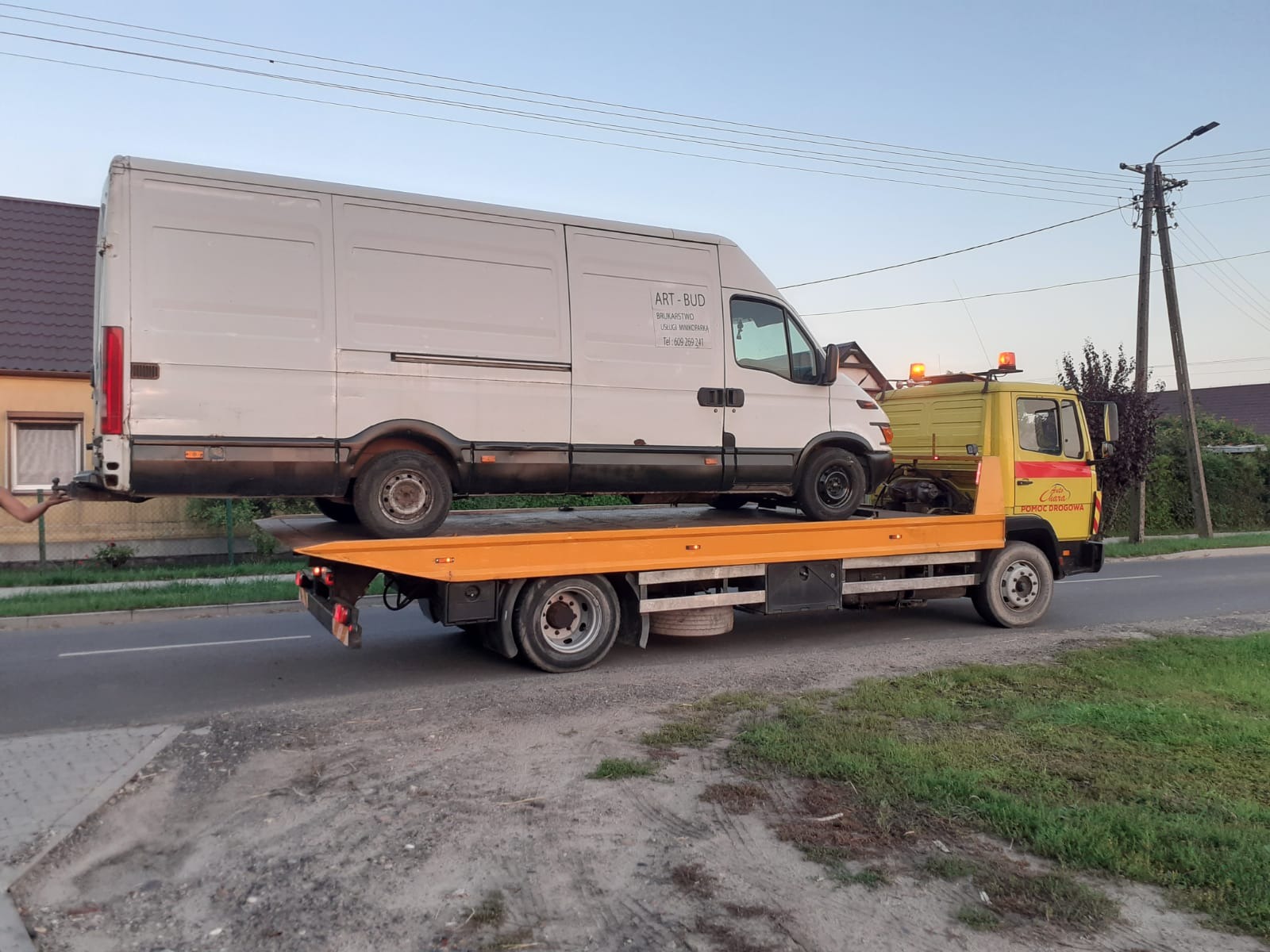 Żółta laweta z włączonymi światłami ostrzegawczymi przewozi białego busa firmy Art-Bud na pomarańczowej platformie, widoczne zabudowania w tle.