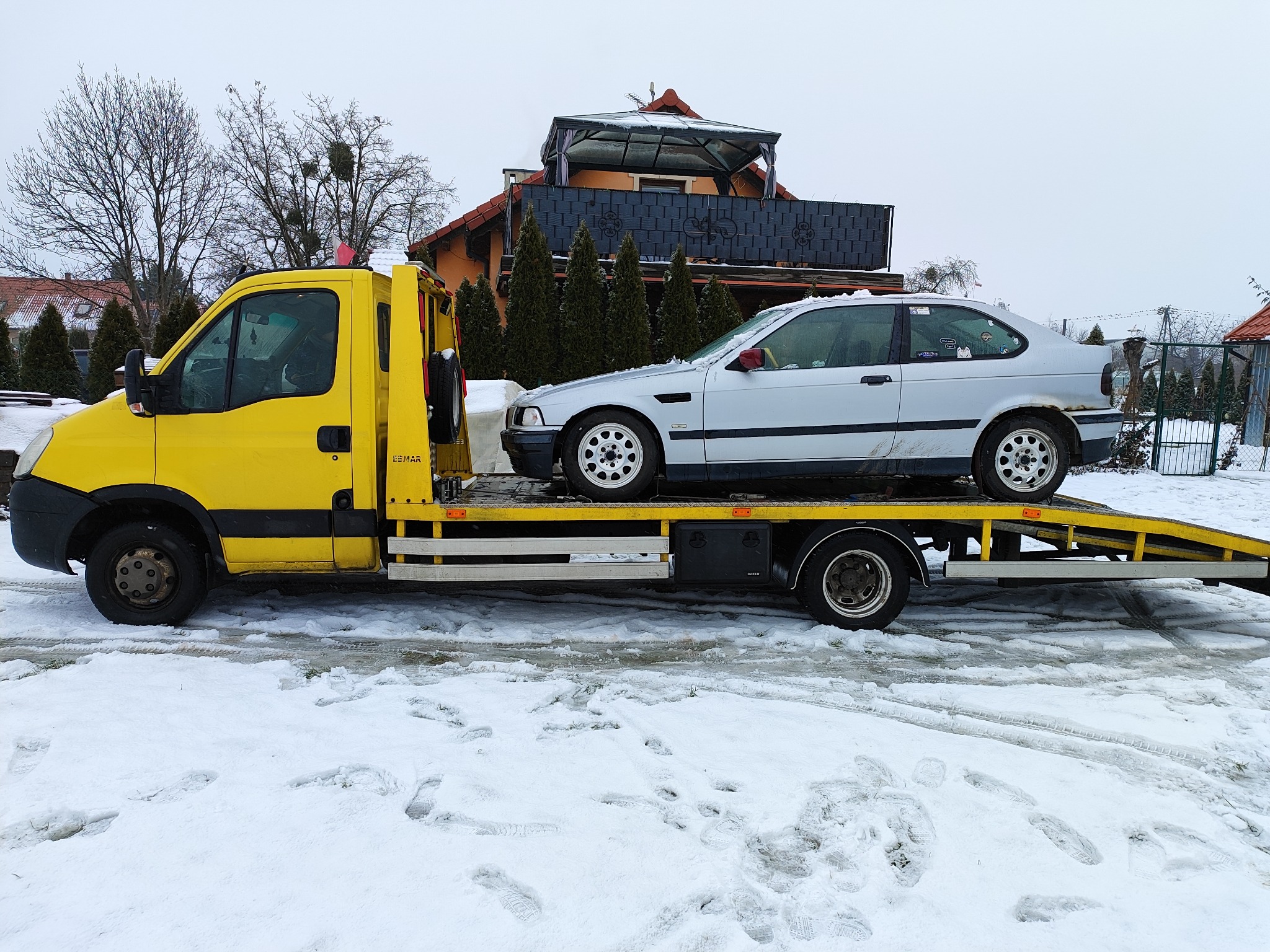 Żółta autolaweta z srebrnym samochodem osobowym na platformie, zimowa sceneria z zśnieżonym podłożem i domem w tle.