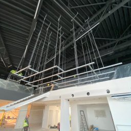 Wisti Krystian Bayger - Montaż konstrukcji sufitowej z metalowych profili pod instalacje w dużej hali, widoczni pracownicy w odblaskowych kamizelkach na podnośniku, surowy stan wykończenia wnętrza.