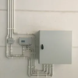 Szara skrzynka elektryczna z czarnym uchwytem zamontowana na białej ścianie, z podłączonymi białymi rurkami instalacyjnymi, widoczne także dwa mniejsze urządzenia elektryczne połączone rurkami.