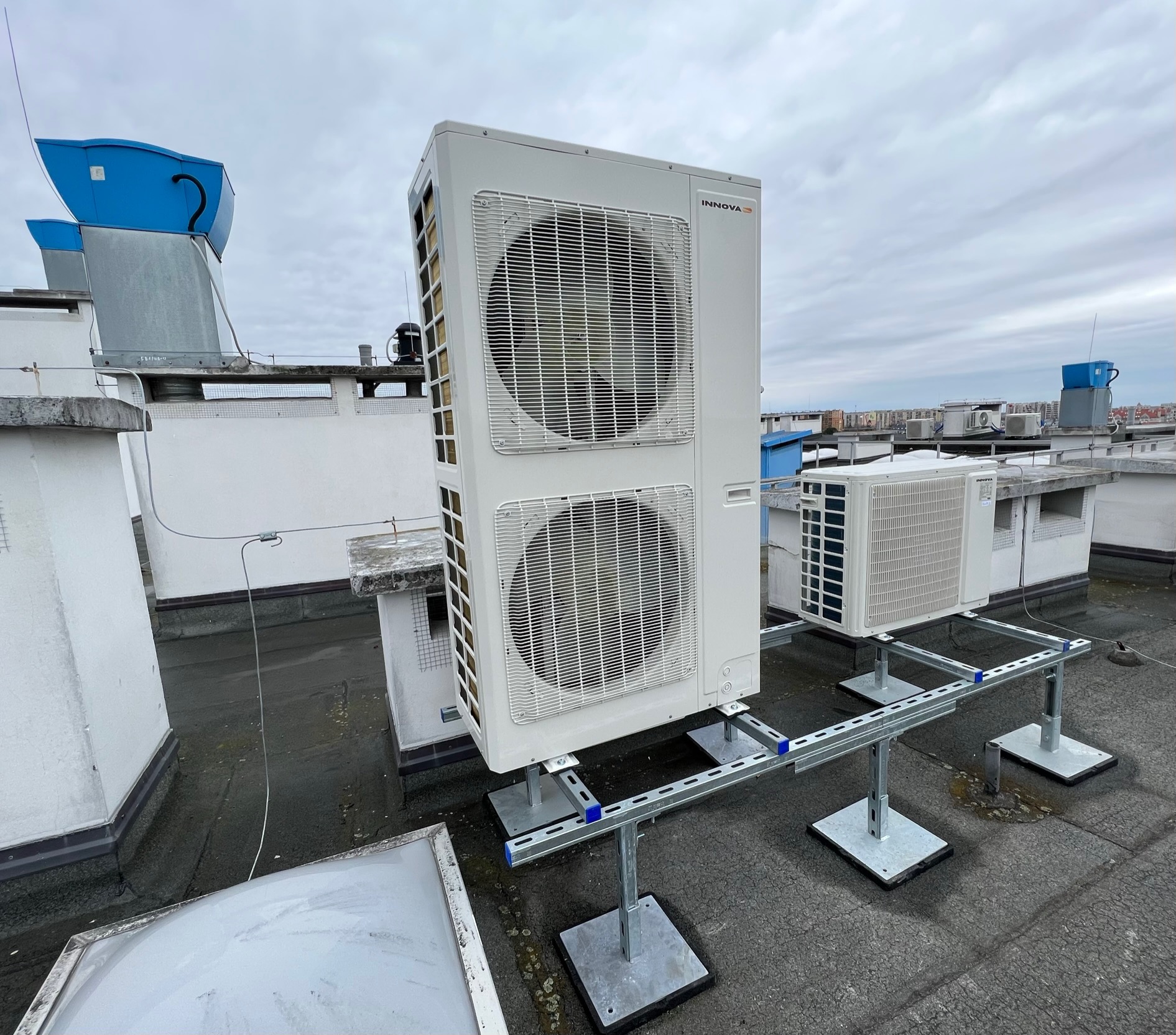 Dwie jednostki zewnętrzne klimatyzacji marki Innova, zamontowane na dachu budynku, na metalowej konstrukcji wsporczej z szarymi podstawami i niebieskimi elementami, widok z niskiej perspektywy.