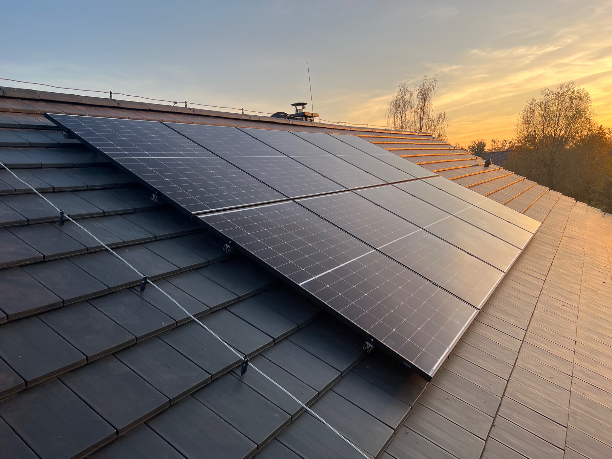 Panel słoneczny zamontowany na ciemnej, ceramicznej dachówce, uchwycony pod kątem o zmierzchu, z widocznymi elementami mocującymi i odgromowymi.