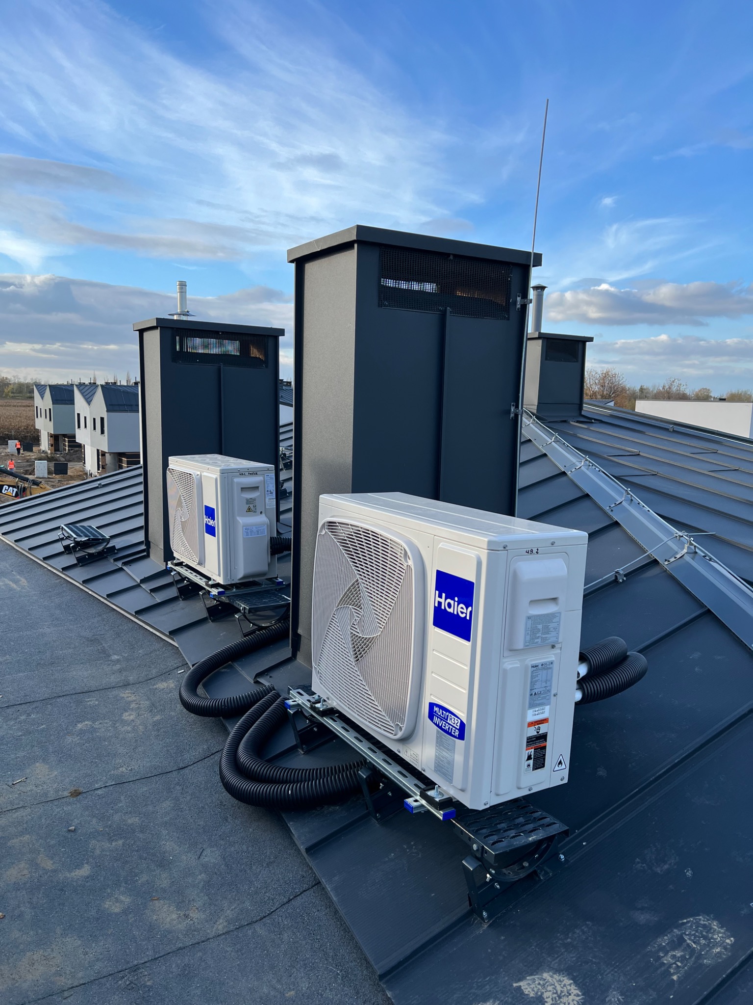 Dwa klimatyzatory marki Haier zamontowane na dachu pokrytym blachą, z widocznymi czarnymi przewodami i elementami montażowymi, na tle błękitnego nieba i zabudowy w oddali.