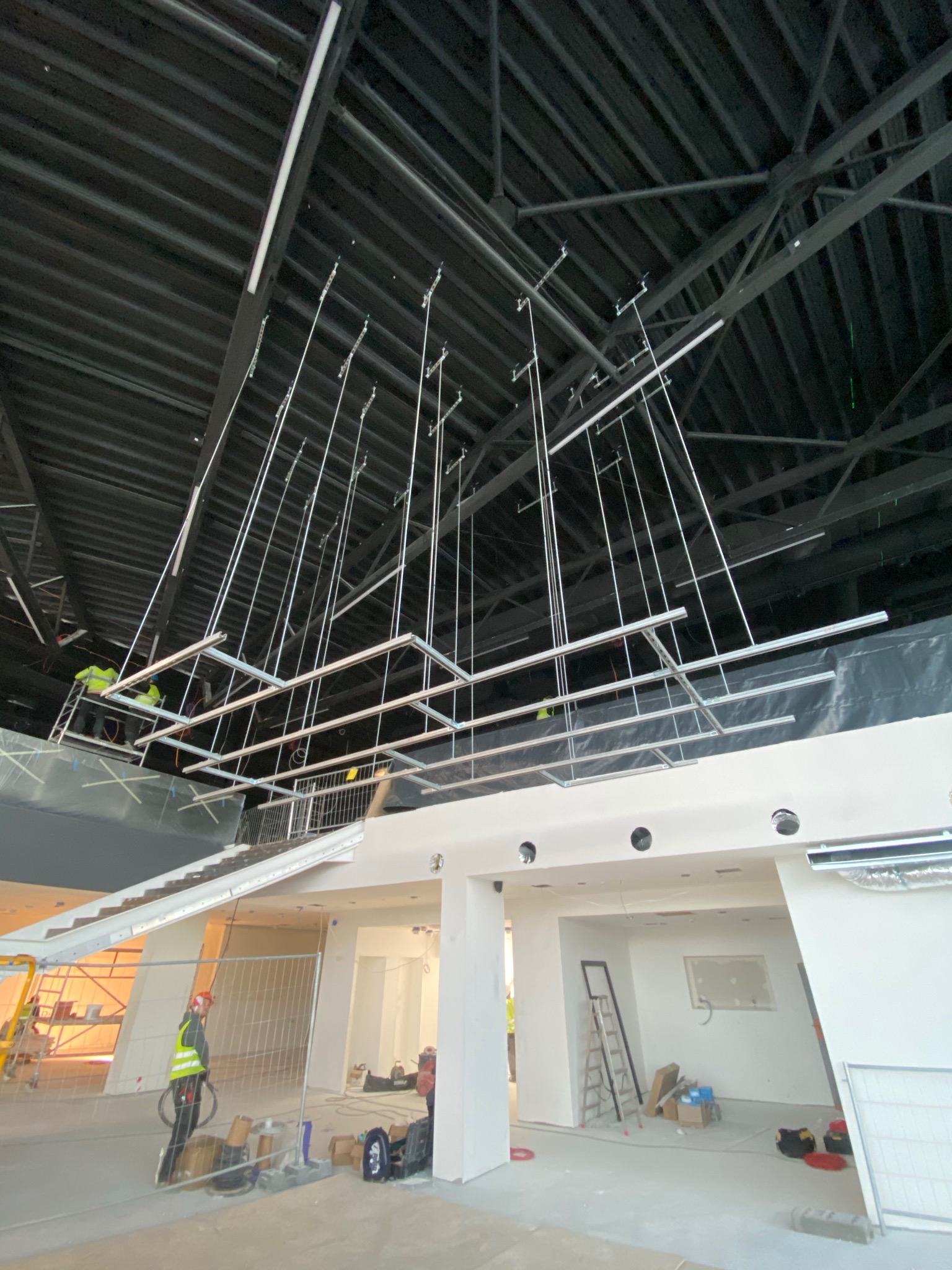 Montaż konstrukcji sufitowej z metalowych profili pod instalacje w dużej hali, widoczni pracownicy w odblaskowych kamizelkach na podnośniku, surowy stan wykończenia wnętrza.