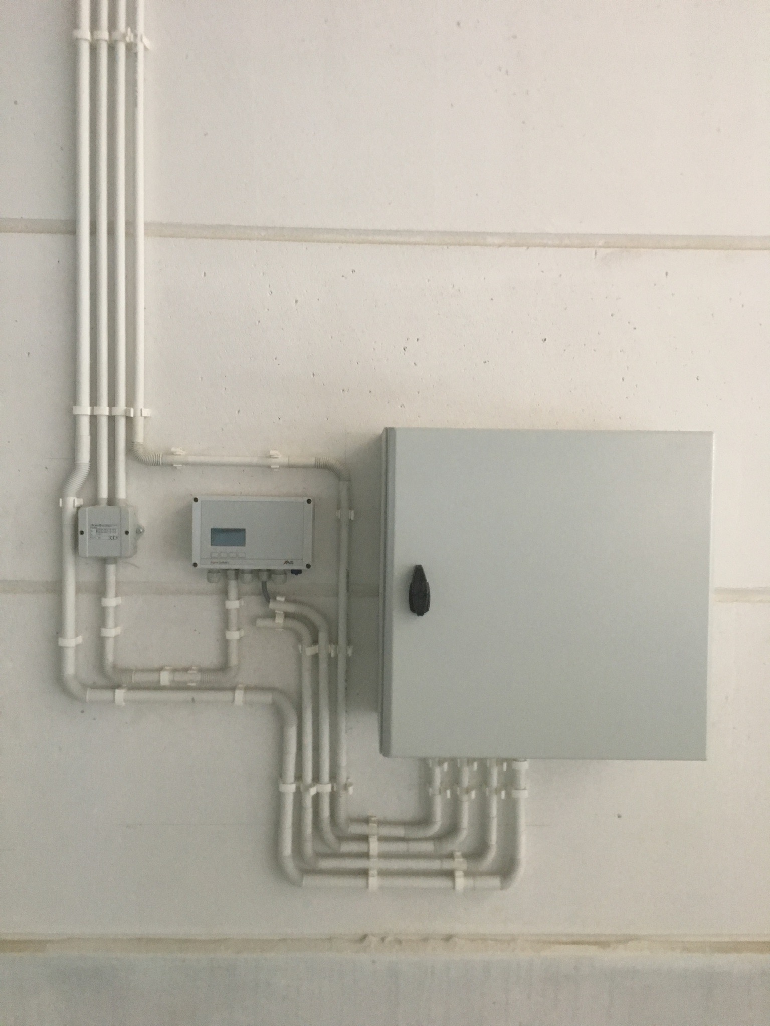 Szara skrzynka elektryczna z czarnym uchwytem zamontowana na białej ścianie, z podłączonymi białymi rurkami instalacyjnymi, widoczne także dwa mniejsze urządzenia elektryczne połączone rurkami.