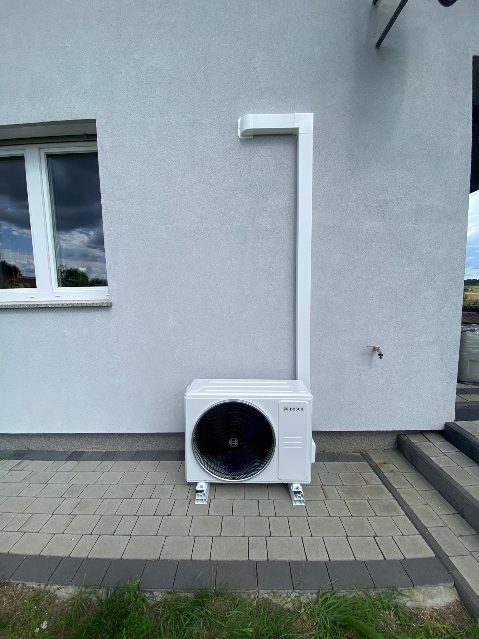 Zewnętrzna jednostka klimatyzacji Bosch zamontowana na elewacji domu z szarym tynkiem, widoczne białe rury odprowadzające skropliny i okno z odbiciem chmur.