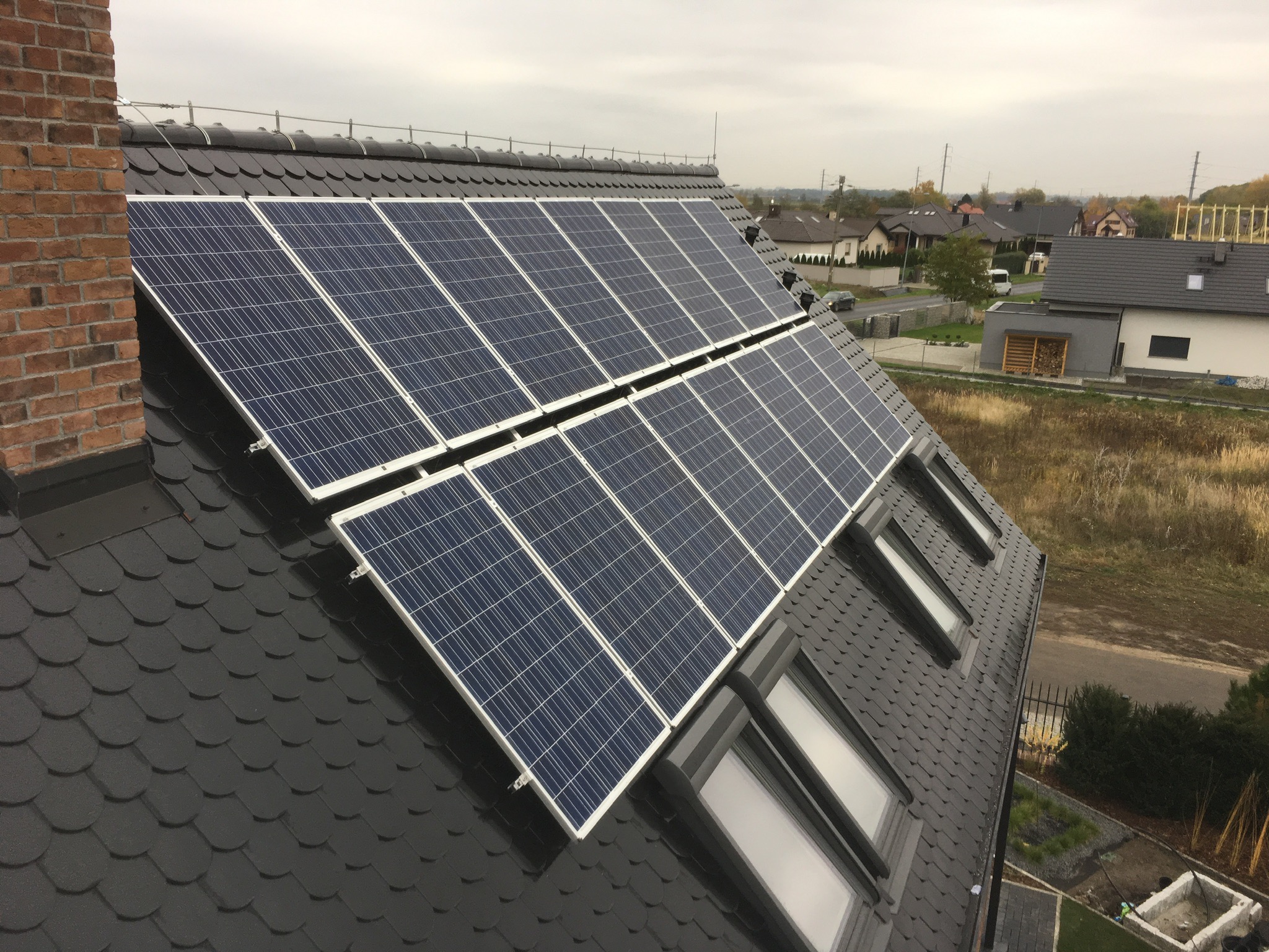 Panele słoneczne zamontowane na dachu domu z dachówką bitumiczną, widoczne okna dachowe i komin z cegły, w tle zabudowa jednorodzinna.