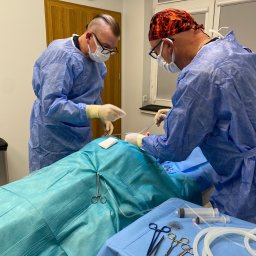 Medi Medic Centrum Medyczne - Dwóch chirurgów w niebieskich strojach operacyjnych, jeden w czepku z płomieniami, przeprowadza zabieg na pacjencie leżącym na stole operacyjnym, widoczne narzędzia chirurgiczne i strzykawki.