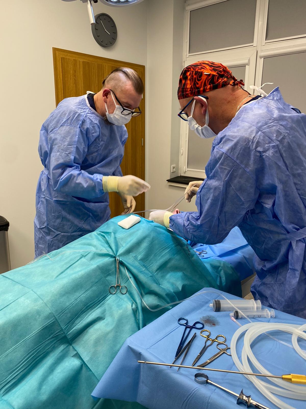 Dwóch chirurgów w niebieskich strojach operacyjnych, jeden w czepku z płomieniami, przeprowadza zabieg na pacjencie leżącym na stole operacyjnym, widoczne narzędzia chirurgiczne i strzykawki.