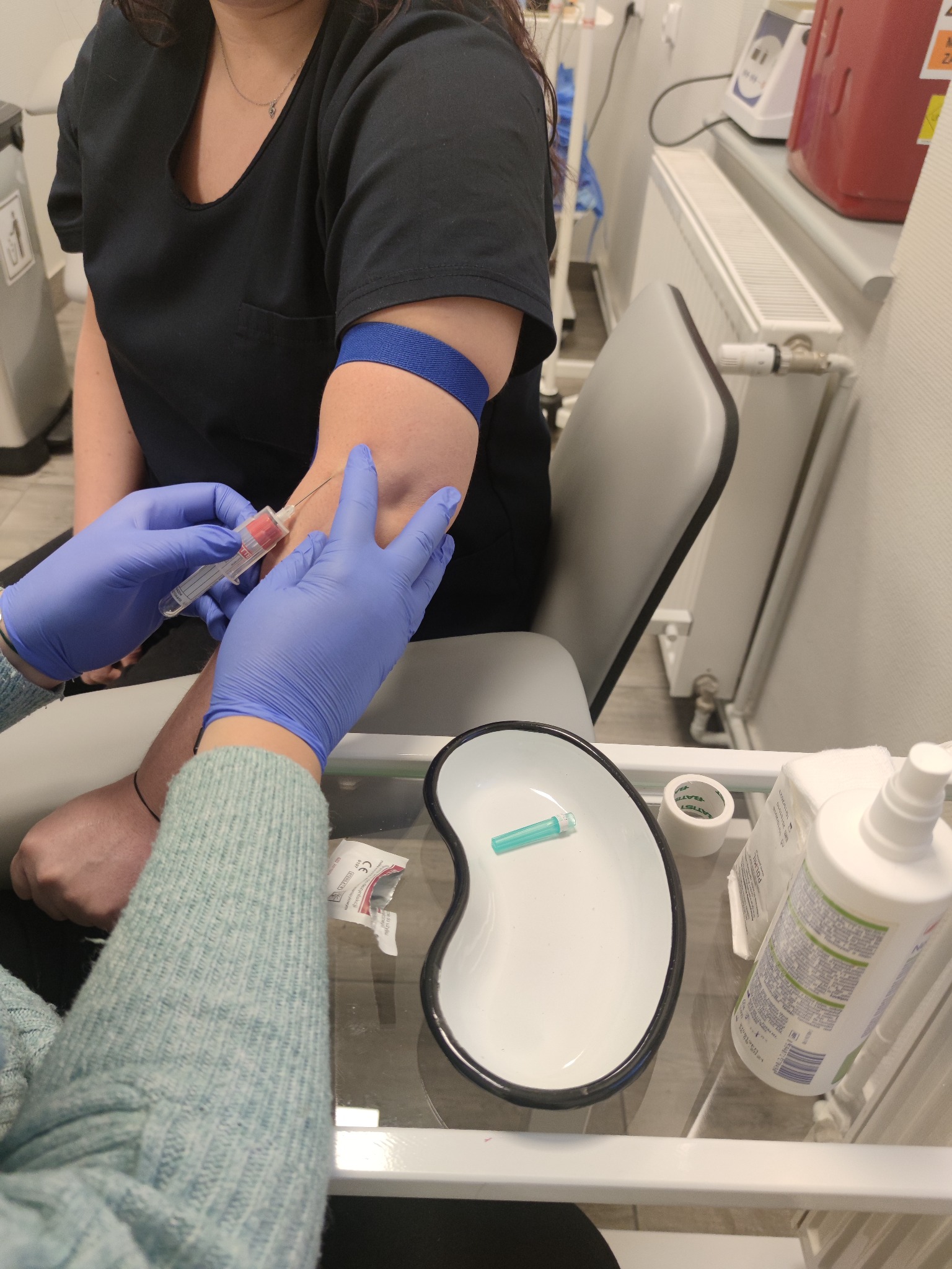 Pobieranie krwi do probówki próżniowej; na pierwszym planie igła, niebieska opaska uciskowa na ramieniu pacjenta, dłonie w niebieskich rękawiczkach, tacka nerkowata z akcesoriami medycznymi.