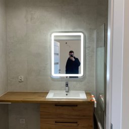 MK INVEST-BUD SPÓŁKA Z OGRANICZONĄ ODPOWIEDZIALNOŚCIĄ - Wykończona łazienka z drewnianą szafką pod umywalką, podświetlanym lustrem i szklanym parawanem nawannowym, widoczny instalator robiący zdjęcie w lustrze.