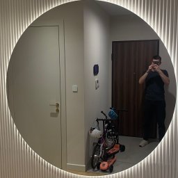 MK INVEST-BUD SPÓŁKA Z OGRANICZONĄ ODPOWIEDZIALNOŚCIĄ - Okrągłe lustro z podświetleniem LED zamontowane na ścianie z dekoracyjnymi panelami pionowymi, odbijające wnętrze przedpokoju z rowerkiem dziecięcym i osobą robiącą zdjęcie.