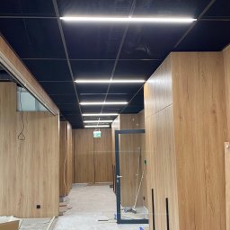 MK INVEST-BUD SPÓŁKA Z OGRANICZONĄ ODPOWIEDZIALNOŚCIĄ - Nowoczesny korytarz biurowy w Warszawie z czarnym sufitem rastrowym, liniowymi lampami LED i jasnymi panelami ściennymi imitującymi drewno, w trakcie wykańczania.