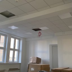 MK INVEST-BUD SPÓŁKA Z OGRANICZONĄ ODPOWIEDZIALNOŚCIĄ - Wykańczane pomieszczenie biurowe w Warszawie z modułowym sufitem podwieszanym, gdzie z otworów zwisają przewody elektryczne. Na podłodze folia, a w tle liczne kartony i krzesła biurowe w folii...