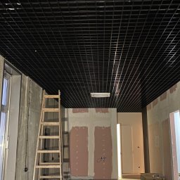 MK INVEST-BUD SPÓŁKA Z OGRANICZONĄ ODPOWIEDZIALNOŚCIĄ - Przestronne wnętrze w fazie wykończenia z czarnym sufitem rastrowym, widocznymi instalacjami, drewnianą drabiną i częściowo otynkowanymi ścianami.