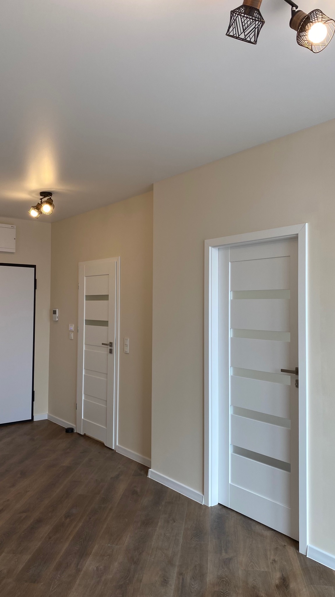 Wykończone wnętrze: Dwa białe, nowoczesne drzwi z przeszkleniami na tle beżowych ścian i drewnopodobnej podłogi, oświetlone lampami sufitowymi.