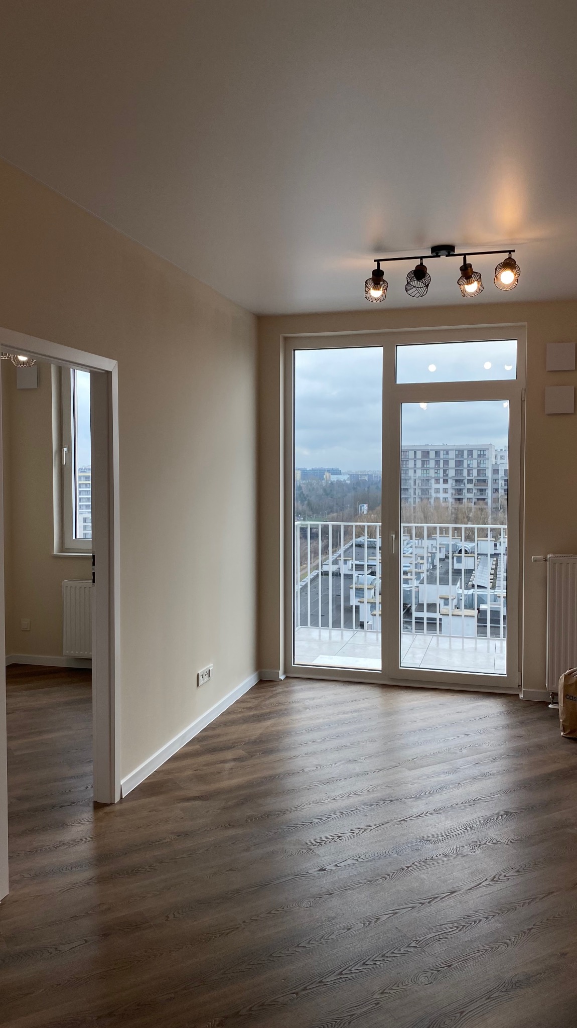 Pusty, jasny pokój z brązowymi panelami podłogowymi i dużym oknem balkonowym z widokiem na miasto, świeżo pomalowane ściany w odcieniach beżu i czarna lampa sufitowa z czterema żarówkami.