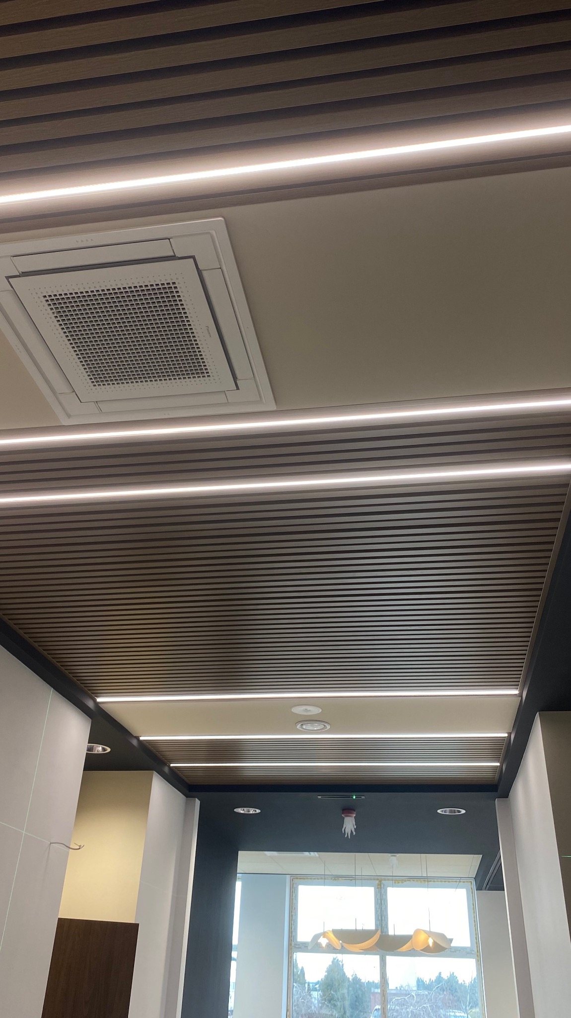 Sufit podwieszany z lamelami i liniowym oświetleniem LED, widoczny klimatyzator kasetonowy i wystrój wnętrza biura.
