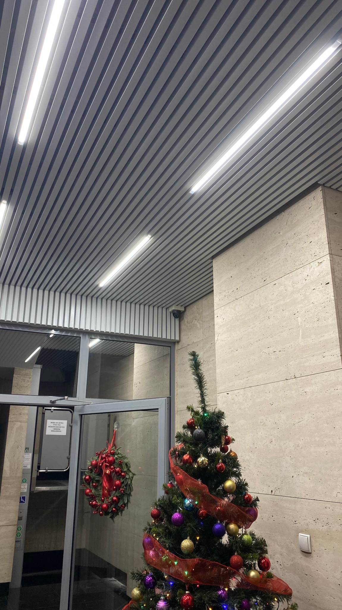 Ozdobiona choinka w holu budynku z podwieszanym sufitem rastrowym i lampami liniowymi.