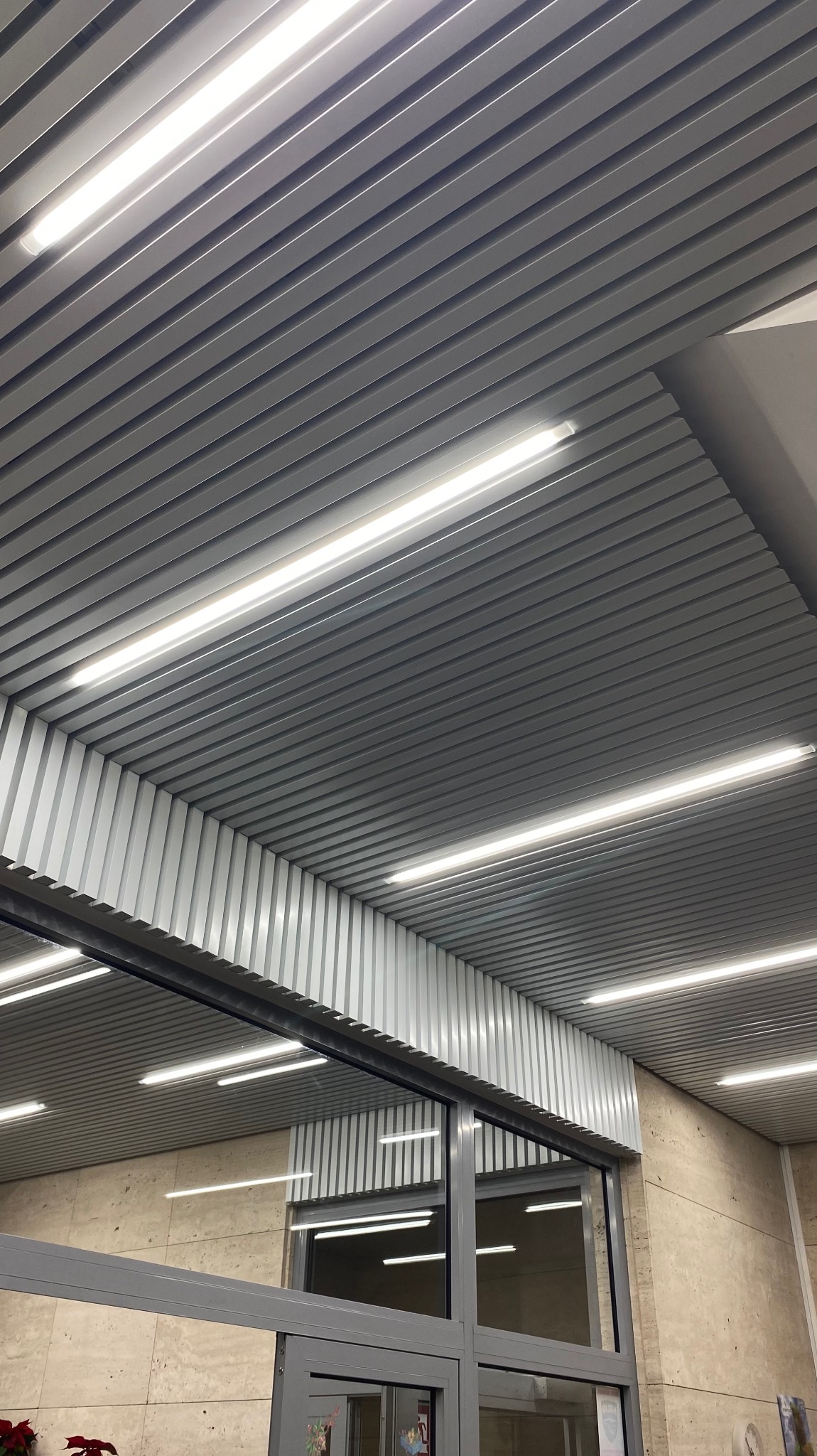 Sufit podwieszany z metalowych paneli z oświetleniem liniowym, widok z dołu pod kątem, fragment przeszklonej ściany z szarymi ramami widoczny w tle.