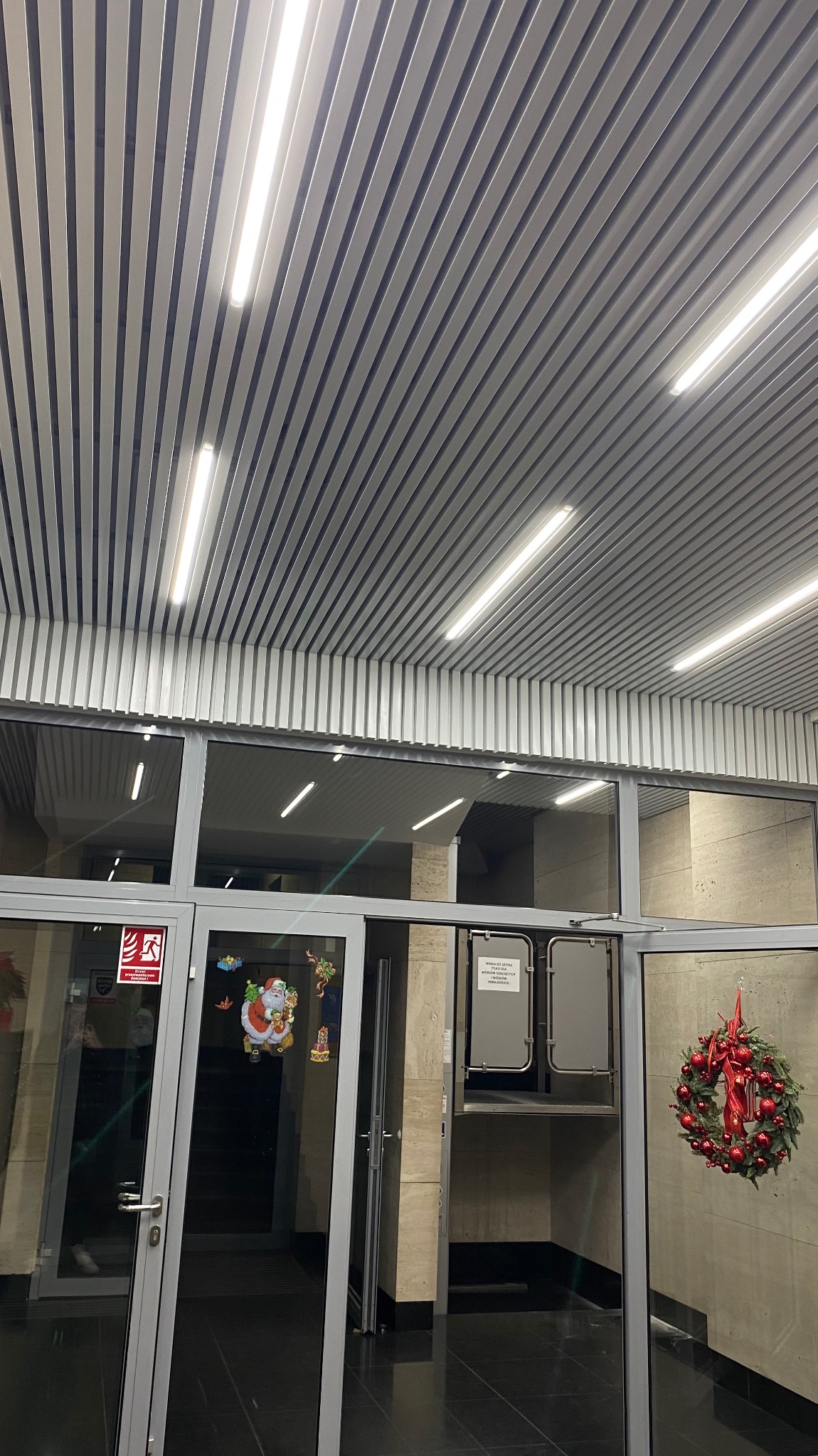 Sufit z metalowych paneli rastrowych z wbudowanym oświetleniem liniowym, widok z dołu, fragment przeszklonego wejścia z dekoracjami świątecznymi.