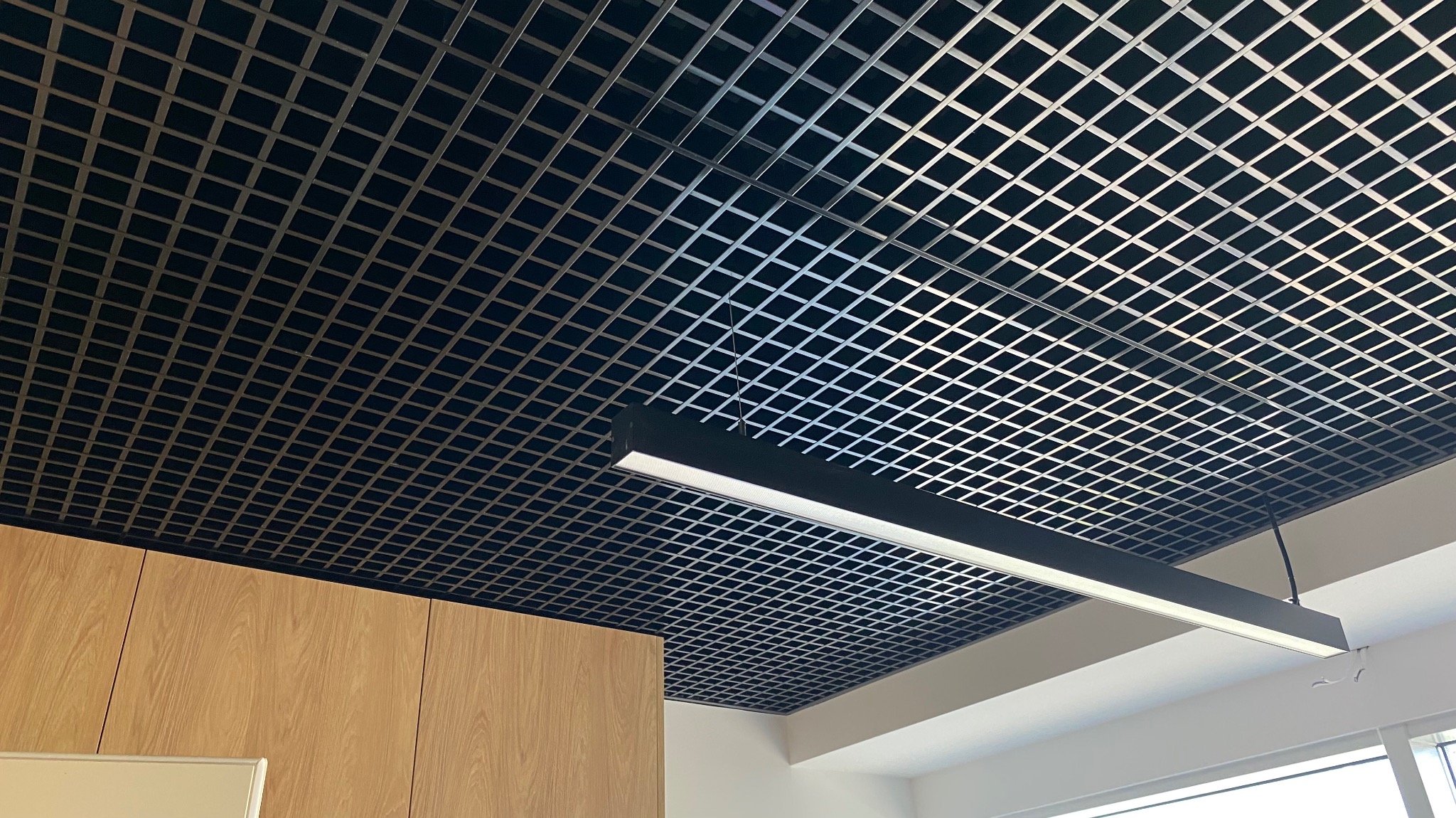 Sufit podwieszany z metalowej siatki w biurze, oświetlony podłużną lampą LED, z widocznymi fragmentami drewnianej zabudowy i okna.