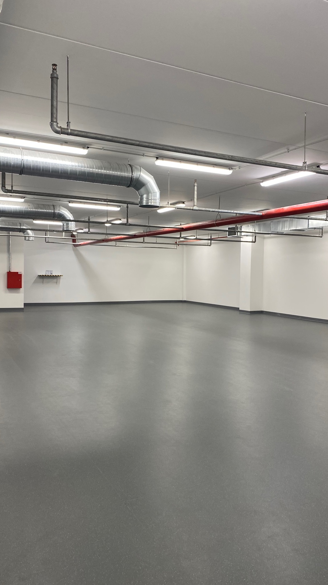 Pusta, jasna hala z szarą podłogą, białymi ścianami i widocznymi instalacjami pod sufitem, w tym rury wentylacyjne i przeciwpożarowe.