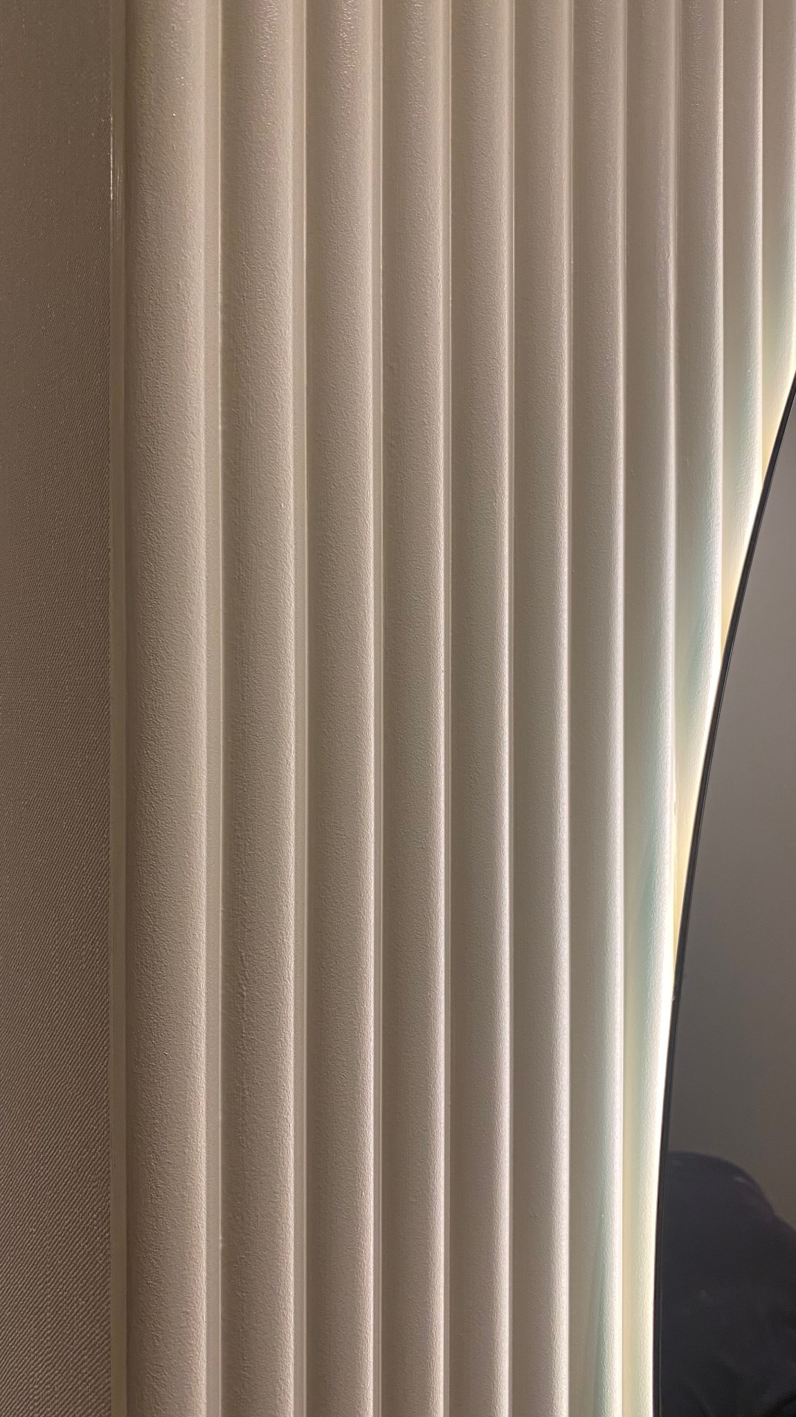 Teksturowana ściana z pionowymi wypukłościami w odcieniach bieli i beżu, oświetlona z boku, z fragmentem ciemnego, błyszczącego elementu (prawdopodobnie lustra lub telewizora) po prawej stronie.