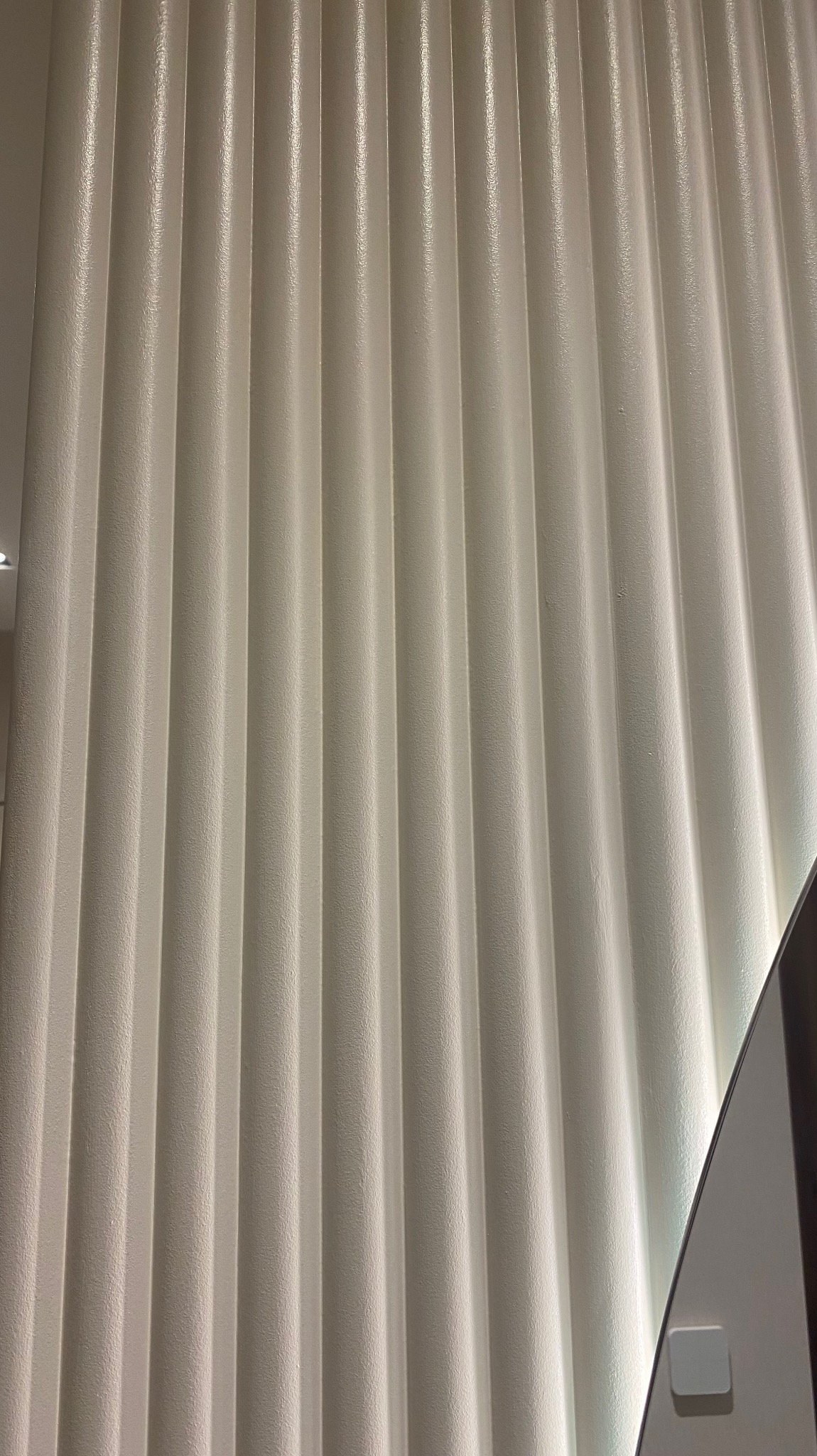 Biała, pionowa ściana z płyty kartonowo-gipsowej, z wyraźnymi, regularnymi żłobieniami, oświetlona z prawej strony, fragment urządzenia widoczny w prawym dolnym rogu.